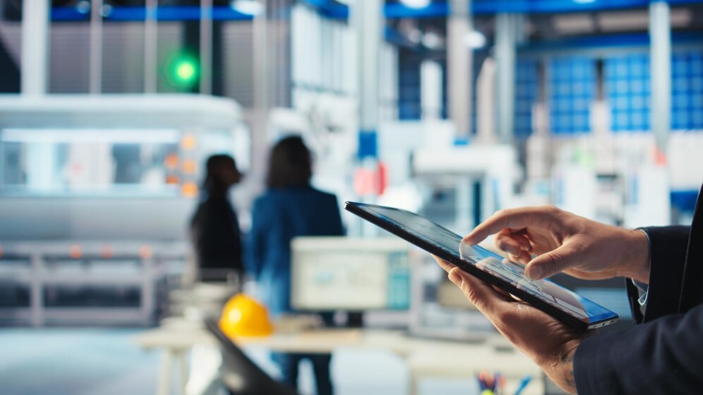 V ospredju roka delavca, ki tipka po tablici, v ozadju dve osebi ob tovarniških strojih