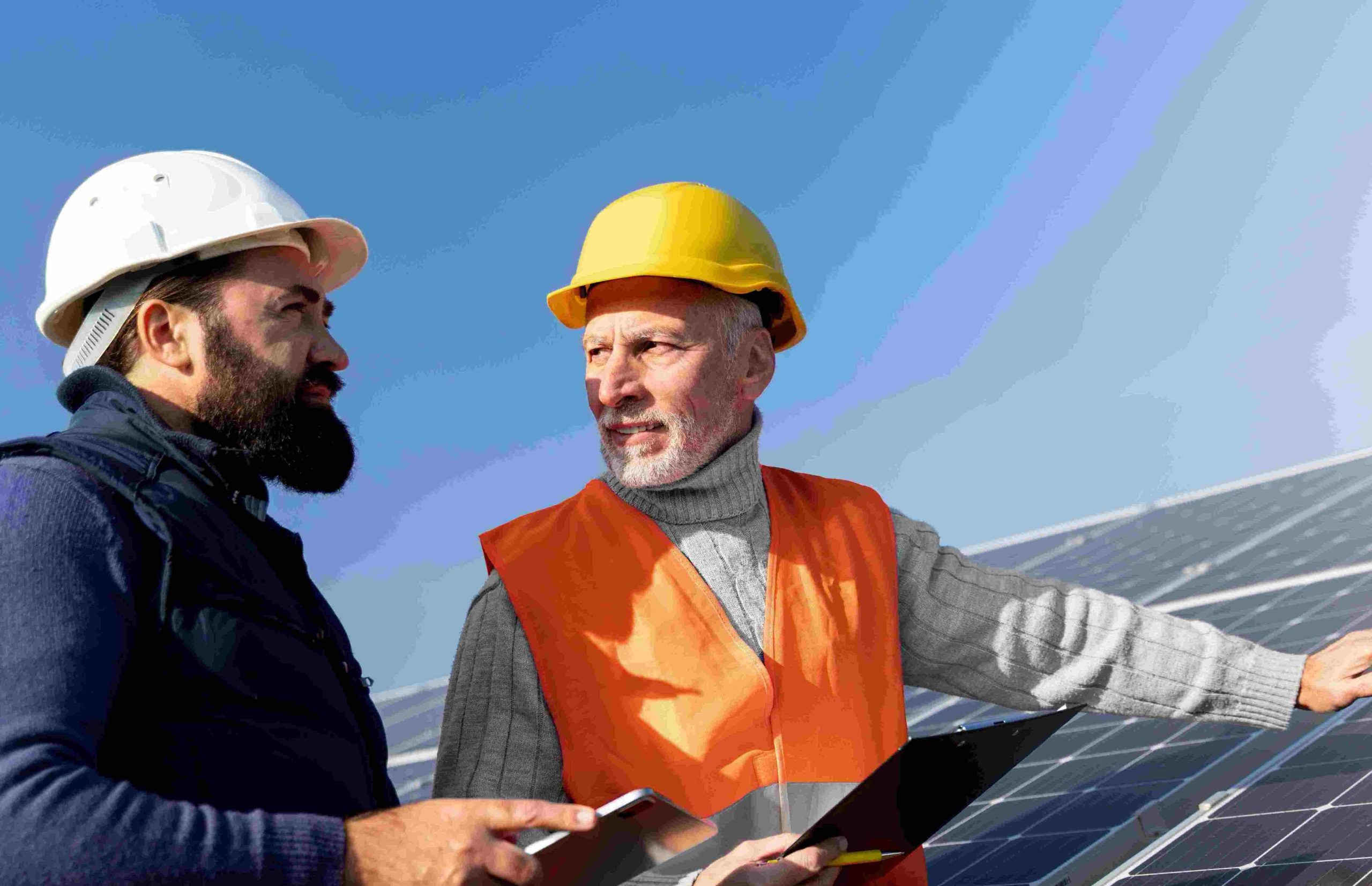 V ospredju na desni strani fotografije moška z delovnima varsnotnima čeladam na glavi v pogovoru, v ozadju vidne sončne celice na strehi.