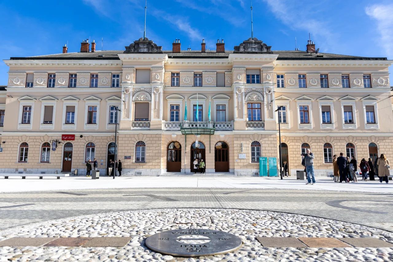 V ospredju na tleh okrogel kovinski znak na meji med Slovenijo in Italijo na Trgu Evropa. V ozadju železniška postaja Nova Gorica.