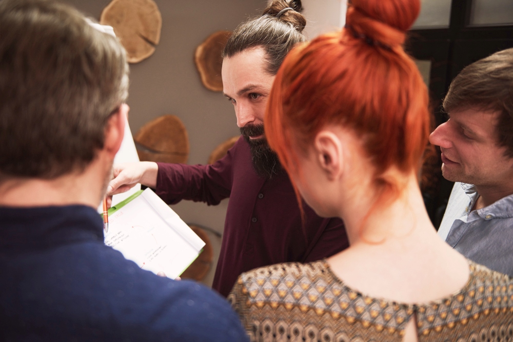 Skupna mlajših ljudi posluša moškega, ki ob stojalu s papirji razlaga napisano. 